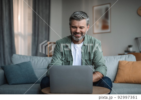 Cheerful retired european male with beard blogger typing on computer or watching video in living room Cheerful retired european male with beard blogger typing on computer or watching video in living room 92997361