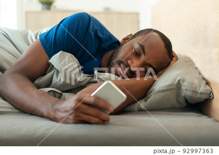 African American Guy Using Cellphone Lying In Bed At Home African American Guy Using Cellphone Lying In Bed At Home 92997363