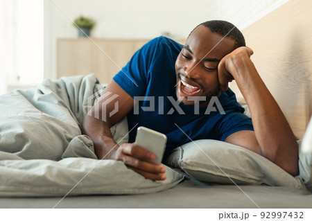 Cheerful Black Man Using Smartphone Lying In Bed In Bedroom Cheerful Black Man Using Smartphone Lying In Bed In Bedroom 92997432
