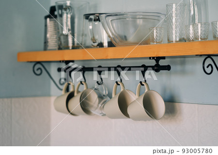 Kitchen open wooden shelf with dishes, glasses and mugs. Mugs are hanging from hooks. Cozy interior in a country house. 93005780