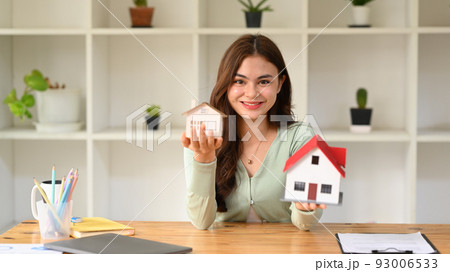 Pretty young woman holding house models and smiling at camera. Real estate, mortgage loan and home insurance concept Pretty young woman holding house models and smiling at camera. Real estate, mortgage loan and home insurance concept 93006533