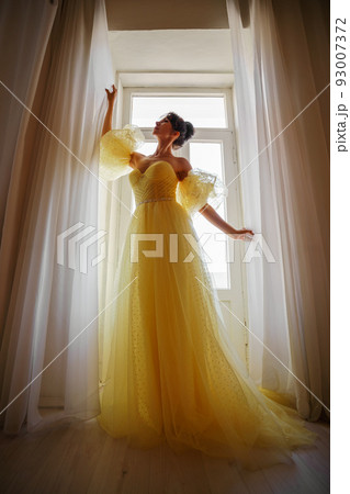 A woman's silhouette in a golden luxurious dress against the background of a window holds a curtain with her hands. Elegant lady in a yellow long silk dress with bare back, back view. 93007372