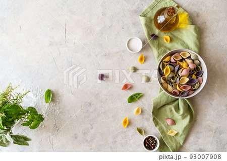 Multi-coloured Italian pasta conchiglie or seashells in bowl, olive oil, spices, herbs, green napkin. Flatly with copy space.  93007908
