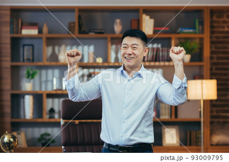 Happy businessman dancing and celebrating victory sitting at a table in a classic office, Asian working at computer Happy businessman dancing and celebrating victory sitting at a table in a classic office, Asian working at computer 93009795