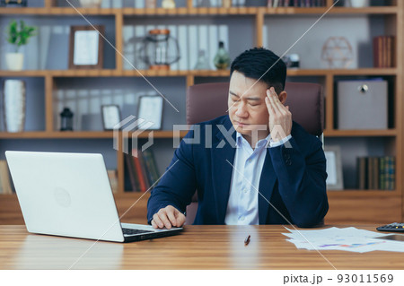 Tired young asian man businessman, worker, lawyer at work. He has a headache, keeps his hands on his head, sits at a desk in the office Tired young asian man businessman, worker, lawyer at work. He has a headache, keeps his hands on his head, sits at a desk in the office 93011569