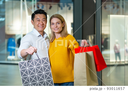 Young asian man and woman couple in supermarket shopping, looking at camera and smiling, holding colored packages with goods 93011657