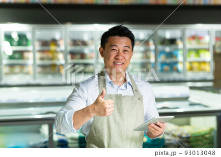 Asian supermarket worker looks into the camera and smiles shows thumbs up, inspects the quality of the goods uses a tablet near the freezers 93013048