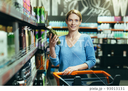 Portrait of a happy woman shopper in the department of household chemicals, woman holding and unwrapping shampoo for hair care, a buyer in a large supermarket Portrait of a happy woman shopper in the department of household chemicals, woman holding and unwrapping shampoo for hair care, a buyer in a large supermarket 93013131