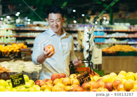 young asian man chooses and picks in eco bag apple fruit or vegetables in the supermarket. male customer standing a grocery store near the counter buys and throws in a reusable package in market young asian man chooses and picks in eco bag apple fruit or vegetables in the supermarket. male customer standing a grocery store near the counter buys and throws in a reusable package in market 93013207