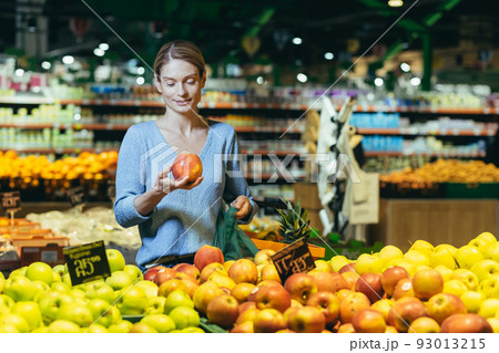 A woman in a supermarket, a buyer chooses an apple fruit, buys and puts an ecological bag in the basket 93013215