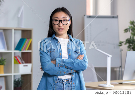 Portrait of a young Asian student girl in glasses and denim clothes, standing, arms crossed, looking at the camera, smiling 93013285