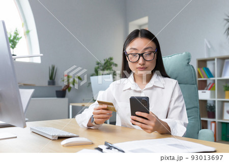 Online shopping. A young beautiful Asian woman holds a mobile phone and a credit card in her hand, makes an order on the phone, pays by card.. Sitting at a table in a modern office. Online shopping. A young beautiful Asian woman holds a mobile phone and a credit card in her hand, makes an order on the phone, pays by card.. Sitting at a table in a modern office. 93013697