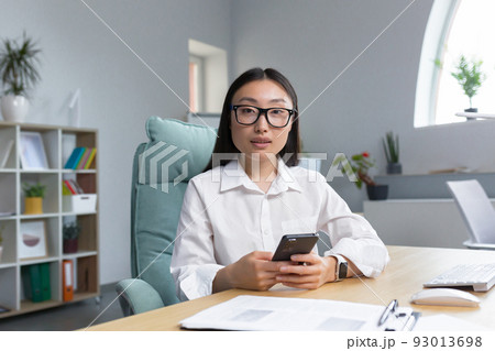 A young beautiful Asian business woman in glasses is holding a phone in her hands, dialing a message. Sitting at a desk in a modern office, looking at the camera. A young beautiful Asian business woman in glasses is holding a phone in her hands, dialing a message. Sitting at a desk in a modern office, looking at the camera. 93013698
