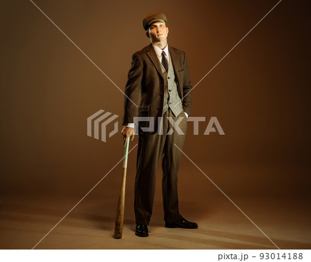 Retro style portrait of young man in image of english gangster, businessman wearing suit and cap standing isolated over dark vintage background. Retro style portrait of young man in image of english gangster, businessman wearing suit and cap standing isolated over dark vintage background. 93014188