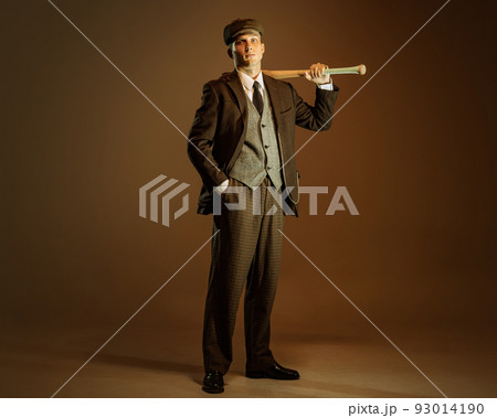 Retro style portrait of young man in image of english gangster, businessman wearing suit and cap standing isolated over dark vintage background. Retro style portrait of young man in image of english gangster, businessman wearing suit and cap standing isolated over dark vintage background. 93014190