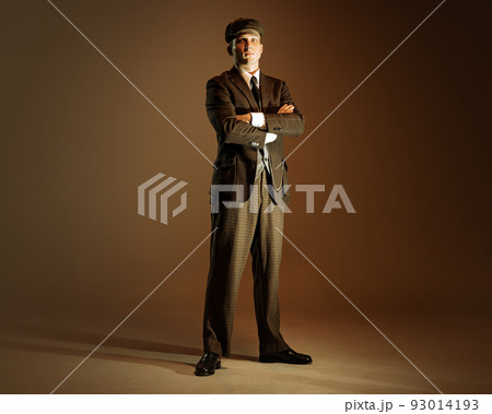 Retro style portrait of young man in image of english gangster, businessman wearing suit and cap standing isolated over dark vintage background. 93014193