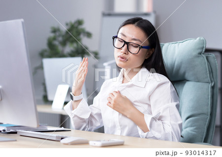 Close-up photo. Heat in the workplace. Young Asian woman sitting in the office at the desk waving her hand at her face, it's very hot, she feels bad, she needs fresh air, take a break. Close-up photo. Heat in the workplace. Young Asian woman sitting in the office at the desk waving her hand at her face, it's very hot, she feels bad, she needs fresh air, take a break. 93014317