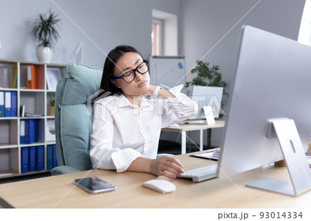 A young Asian female office worker in glasses and a white shirt sits at a desk at a computer, tired, exhausted, holding her hand to her neck, feeling pain. A young Asian female office worker in glasses and a white shirt sits at a desk at a computer, tired, exhausted, holding her hand to her neck, feeling pain. 93014334
