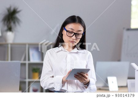 Young beautiful Asian teacher working with tablet, in classroom with computers at school, woman in white shirt and glasses reads from tablet 93014379