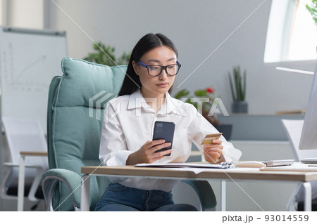 Beautiful and confident Asian business woman working in the office, making banking transactions, using the phone and banking application, holding a credit card, for online shopping. 93014559