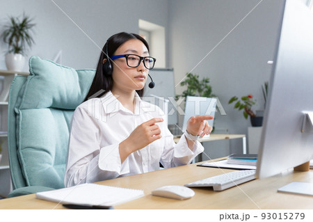 Consultant woman working remotely using headset for video call, talking to customers, helpline, Asian woman working in office. Consultant woman working remotely using headset for video call, talking to customers, helpline, Asian woman working in office. 93015279