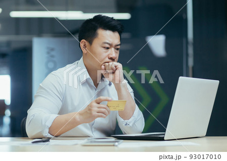 Thoughtful and serious Asian businessman trying to make a bank transaction, man working in office with laptop, holding credit bank card 93017010