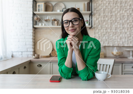 Young beautiful woman in glasses and a green casual shirt, at home in the kitchen, looks at the webcam, video call listens to the interlocutor, view of the webcam Young beautiful woman in glasses and a green casual shirt, at home in the kitchen, looks at the webcam, video call listens to the interlocutor, view of the webcam 93018036