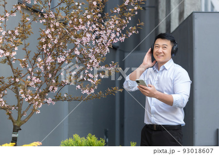 Cheerful and smiling successful Asian business man listening to music using smartphone and big headphones, manager resting outside office in shirt Cheerful and smiling successful Asian business man listening to music using smartphone and big headphones, manager resting outside office in shirt 93018607