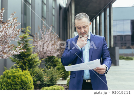 Senior and experienced gray haired boss man near office pensive reading financial report document outside office, businessman thinking Senior and experienced gray haired boss man near office pensive reading financial report document outside office, businessman thinking 93019211