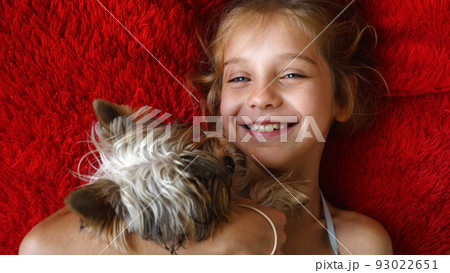 A beautiful blond school-age girl lies on a red pillow with a Yorkshire Terrier dog. The child is smiling with the dog in her arms. A beautiful blond school-age girl lies on a red pillow with a Yorkshire Terrier dog. The child is smiling with the dog in her arms. 93022651