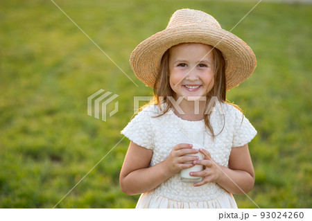 Happy little girl in straw hat drinks glass of milk on lawn Happy little girl in straw hat drinks glass of milk on lawn 93024260