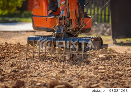A mini excavator with a rake loosens the ground. landscape preparation. A mini excavator with a rake loosens the ground. landscape preparation. 93024268