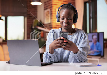 Smiling african american remote worker in headphones typing message on mobile phone. Female freelancer watching videos on smartphone in modern home office with beautiful warm sunset light 93027628