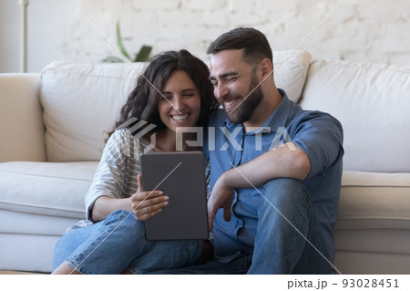 Cheerful couple of satisfied gadget users sitting on floor Cheerful couple of satisfied gadget users sitting on floor 93028451