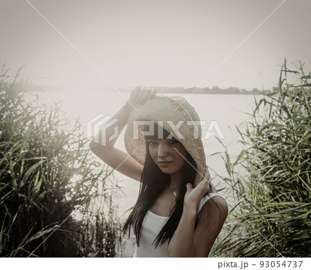 Young woman in vintage hat standing next to riverside with reeds, copyspace on sky 93054737