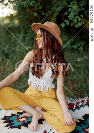 A young hippie woman in eco-clothing has fun laughing and cringing sitting on a plaid outside in the fall with a hat and yellow sunglasses. Travel lifestyle A young hippie woman in eco-clothing has fun laughing and cringing sitting on a plaid outside in the fall with a hat and yellow sunglasses. Travel lifestyle 93076150