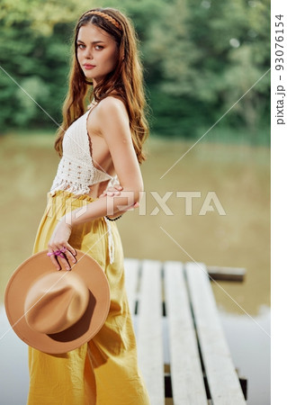 A young woman smiling in an image of a hippie and eco-dress dancing in nature by the lake wearing a hat and yellow pants in the summer sunset A young woman smiling in an image of a hippie and eco-dress dancing in nature by the lake wearing a hat and yellow pants in the summer sunset 93076154