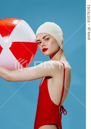Conceptual fashion. Charming girl in a red swimsuit swimming cap with a ball on her face on a blue background Conceptual fashion. Charming girl in a red swimsuit swimming cap with a ball on her face on a blue background 93076161
