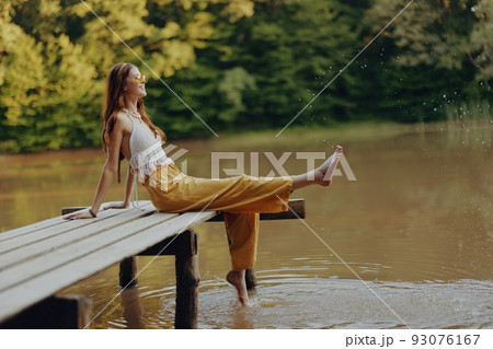 A young happy hippie woman sitting on the lake shore on a bridge in eco clothing made of natural materials in harmony with nature in the fall A young happy hippie woman sitting on the lake shore on a bridge in eco clothing made of natural materials in harmony with nature in the fall 93076167