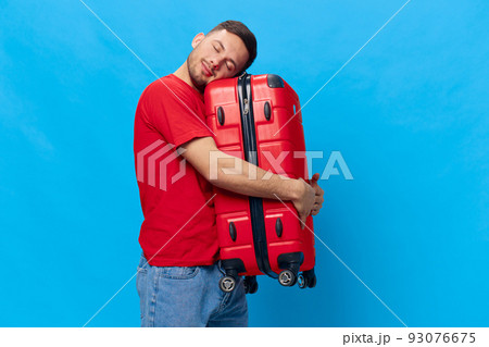 Enjoyed joyful tanned handsome man in red t-shirt hugging with suitcase waiting impatiently for holiday posing isolated on blue studio background. Copy space Banner Mockup. Trip journeys concept 93076675
