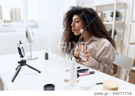 Funny tanned handsome curly Latin beauty blogger in linen shirt teach how to do cheekbones in home white interior. Copy space Mockup Banner. Influencer record video review blog using smartphone 93077758