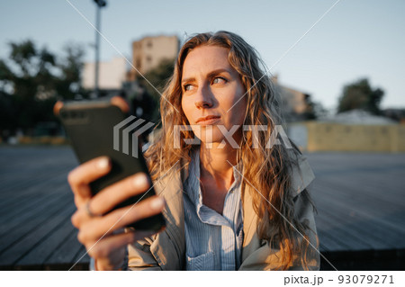 Young casual woman in coat sitting on bench on sea embankment using smartphone Young casual woman in coat sitting on bench on sea embankment using smartphone 93079271