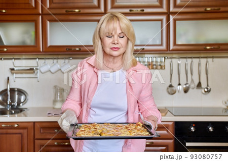 Portrait of mature 50s woman with pan of cooked meat at home in kitchen. Portrait of mature 50s woman with pan of cooked meat at home in kitchen. 93080757