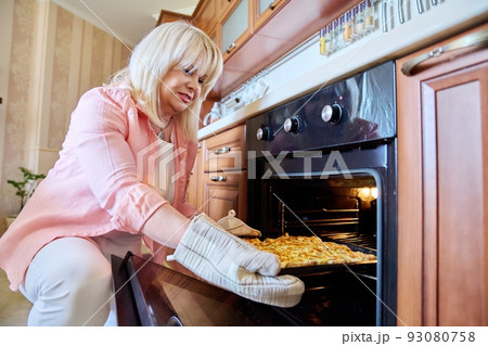 Mature woman cooking meat in oven, at home in kitchen Mature woman cooking meat in oven, at home in kitchen 93080758