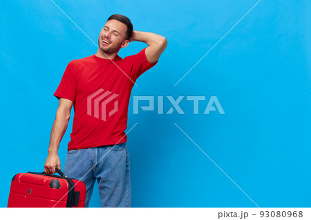 Smiling dreaming tanned handsome man in red t-shirt wait impatiently for vacation hold suitcase posing isolated on blue studio background. Copy space Banner Mockup. Trip journeys concept 93080968