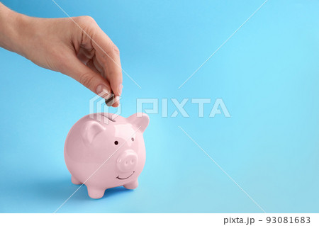 Woman putting coin into piggy bank on light blue background, closeup. Space for text Woman putting coin into piggy bank on light blue background, closeup. Space for text 93081683