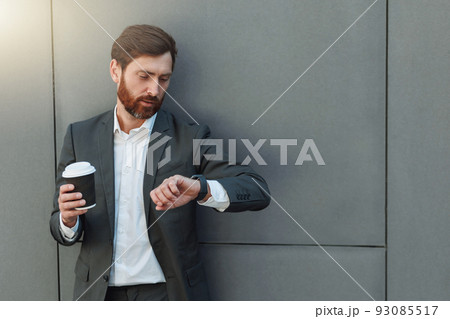 Focused european businessman in suit with coffee watching on his wrist watch during break Focused european businessman in suit with coffee watching on his wrist watch during break 93085517
