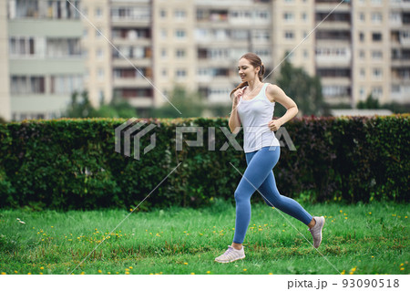 Woman in white t-shirt and leggings takes morning jog in residential area of city next to high-rise apartment building. Woman in white t-shirt and leggings takes morning jog in residential area of city next to high-rise apartment building. 93090518