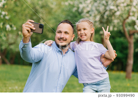 Dad and his six-year-old daughter take selfies on the phone, smiling and having fun on camera in the park in summer or spring 93099490