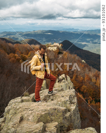 Happy traveler is standing on rock and enjoying the journey. Scenic landscape with view to the autumn forest and mountains. 93101062